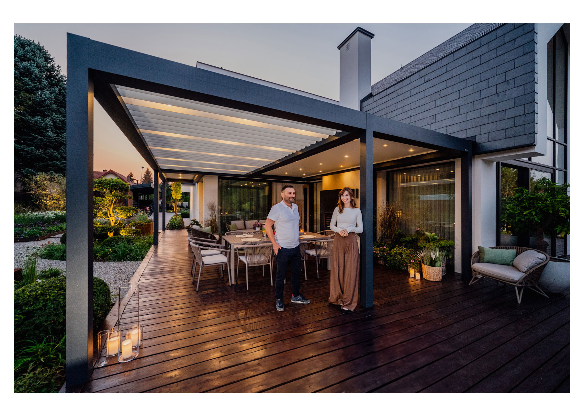 Nowoczesna pergola z regulowanym zadaszeniem nad drewnianym tarasem, oświetlony ogród w tle, para stojąca na tarasie, wieczorny krajobraz.