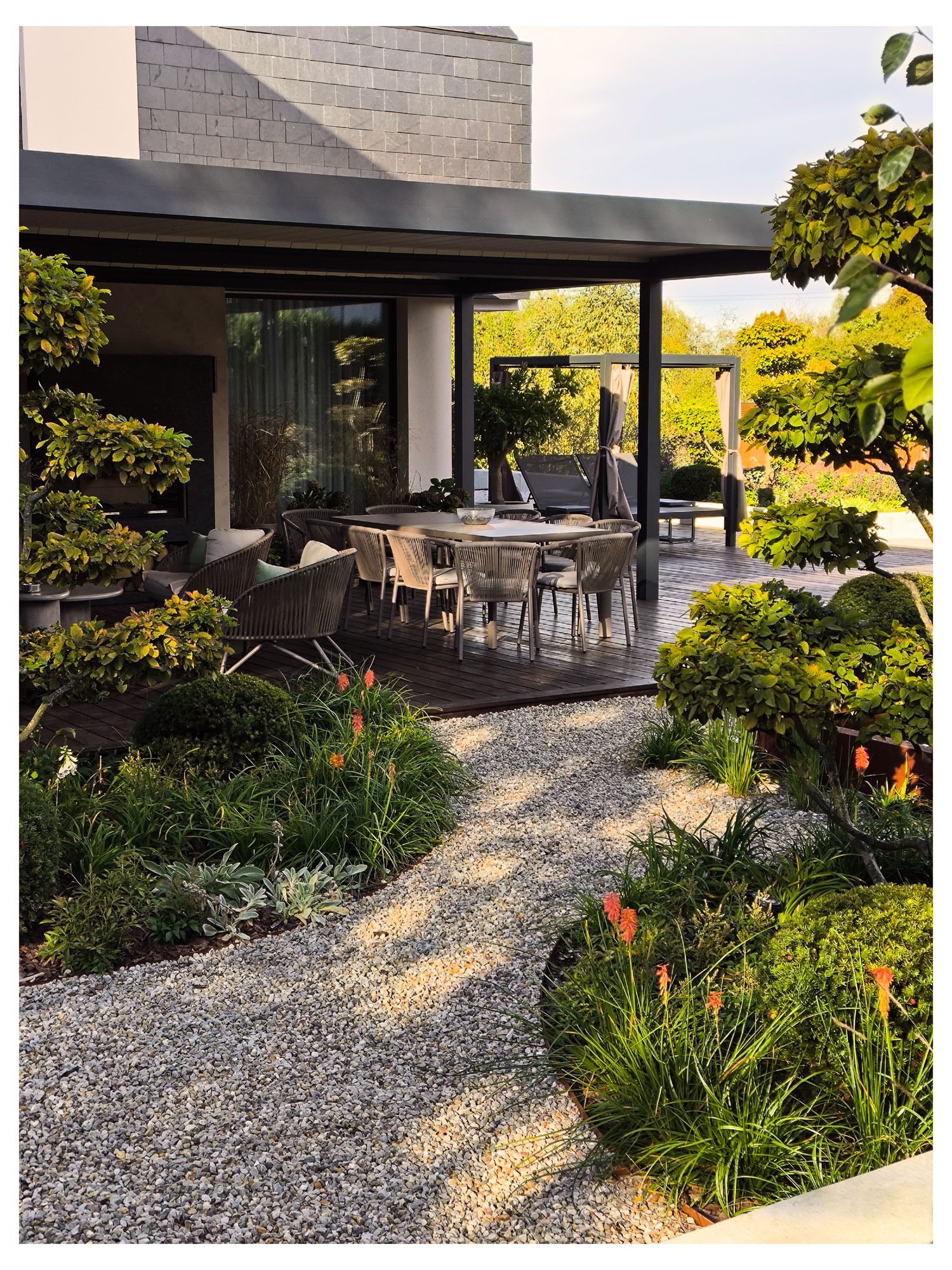 Nowoczesny ogród z tarasem z drewna, ścieżką żwirową i egzotyczną roślinnością. Widoczne meble ogrodowe i pergola. Całość w ciepłej, słonecznej tonacji.