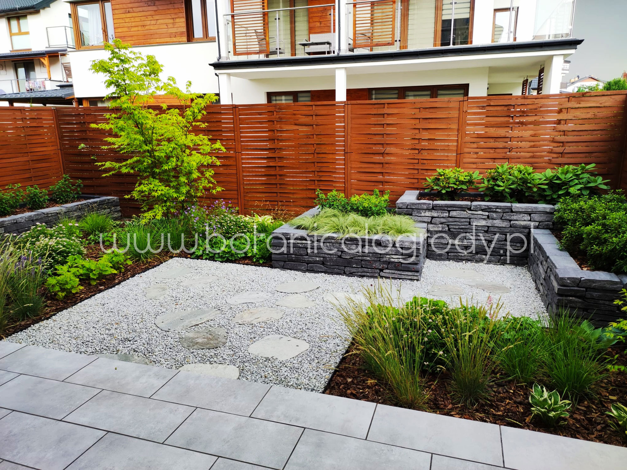 Nowoczesny ogród z kamiennymi rabatami, żwirową ścieżką i drewnianym ogrodzeniem. Widoczne rośliny ozdobne i drzewo. Minimalistyczny design.