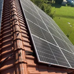 Panele słoneczne zamontowane na dachu z czerwonej dachówki ceramicznej, widoczne kable i mocowania, w tle trawnik i budynki.