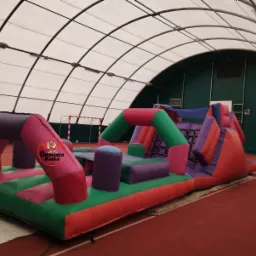 Kolorowy, dmuchany tor przeszkód z logo restauracji w hali sportowej z bramką do piłki nożnej w tle.
