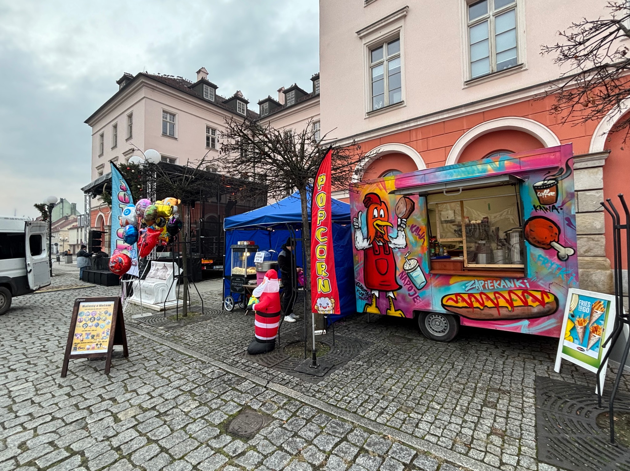 Kolorowy food truck z grafiką kurczaka i zapiekanki, stoisko z popcornem, balony i dmuchany Mikołaj na brukowanym placu miejskim podczas wydarzenia plenerowego.