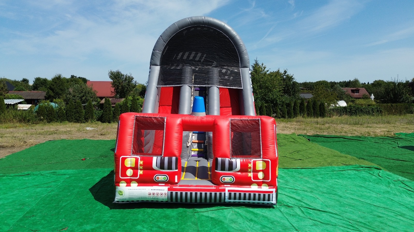 Dmuchany zamek w kształcie wozu strażackiego na zielonej trawie, z widocznymi elementami drabiny i zjeżdżalni, na tle drzew i domów w słoneczny dzień.