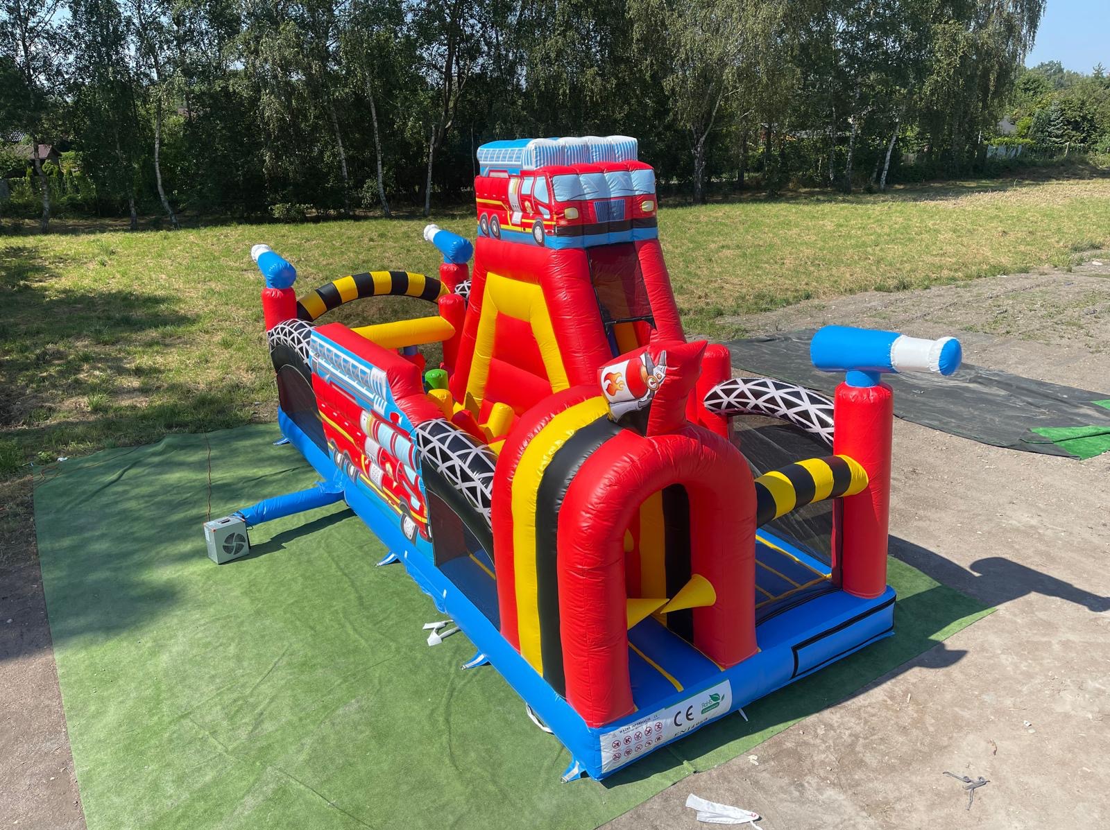 Kolorowy, dmuchany zamek w kształcie wozu strażackiego na zielonej macie, z drzewami w tle. Atrakcja eventowa dla dzieci, zjeżdżalnia i elementy do wspinaczki.