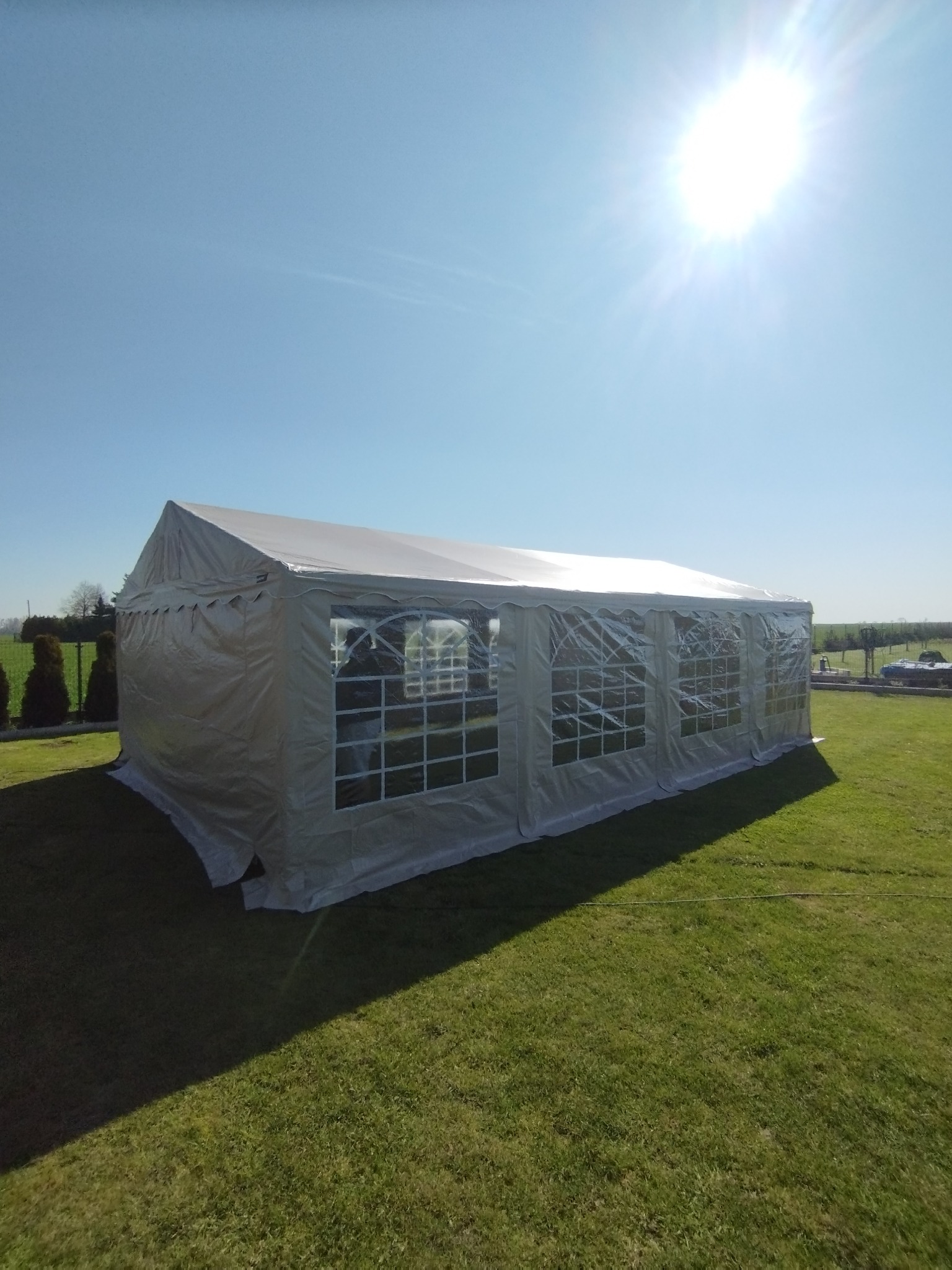 Rozłożony biały namiot eventowy z przezroczystymi oknami na zielonym trawniku w słoneczny dzień.