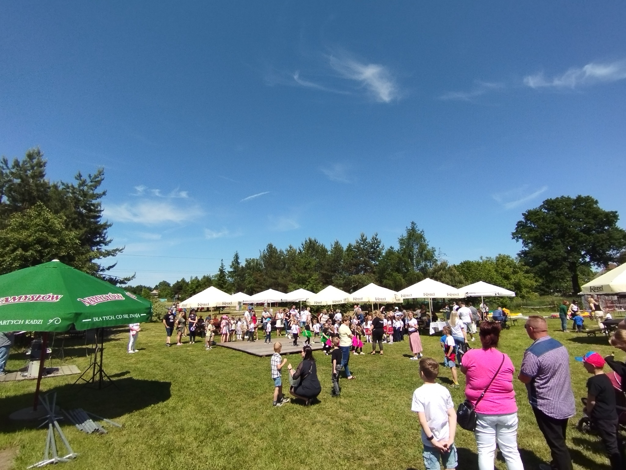 Plenerowa impreza z białymi namiotami, dzieci na scenie, zielony parasol z napisem 'Namysłów', błękitne niebo z delikatnymi chmurami.