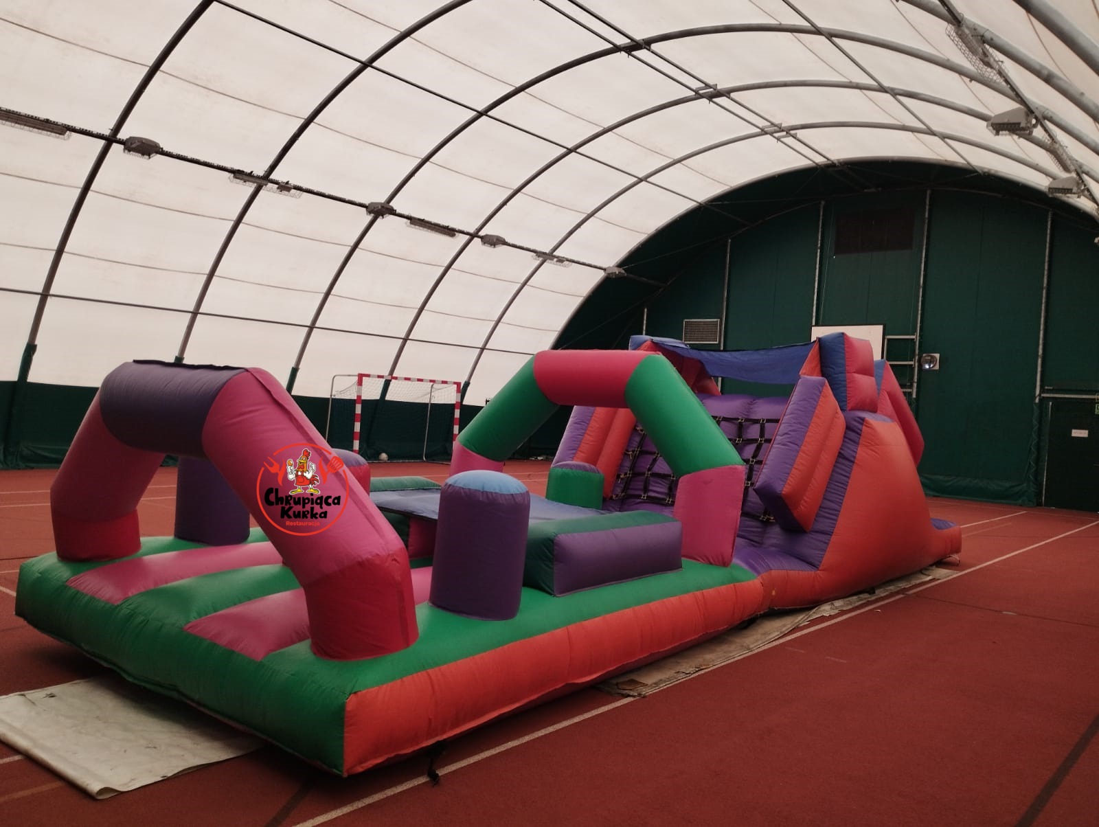 Kolorowy, dmuchany tor przeszkód z logo restauracji w hali sportowej z bramką do piłki nożnej w tle.