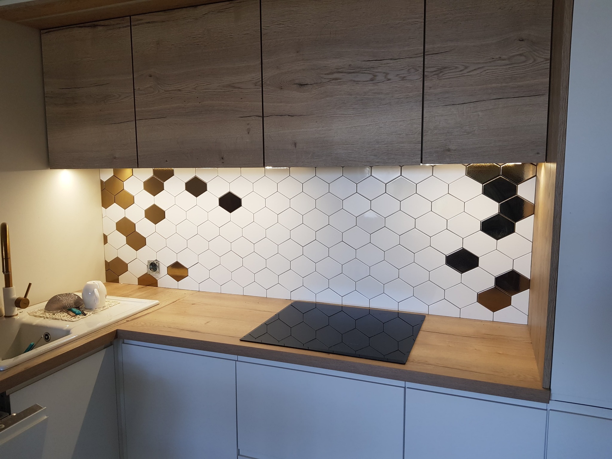 Nowoczesna kuchnia z heksagonalną mozaiką na ścianie, łączącą białe, złote i czarne płytki, drewniane szafki wiszące i blat, oraz indukcyjna płyta grzewcza.