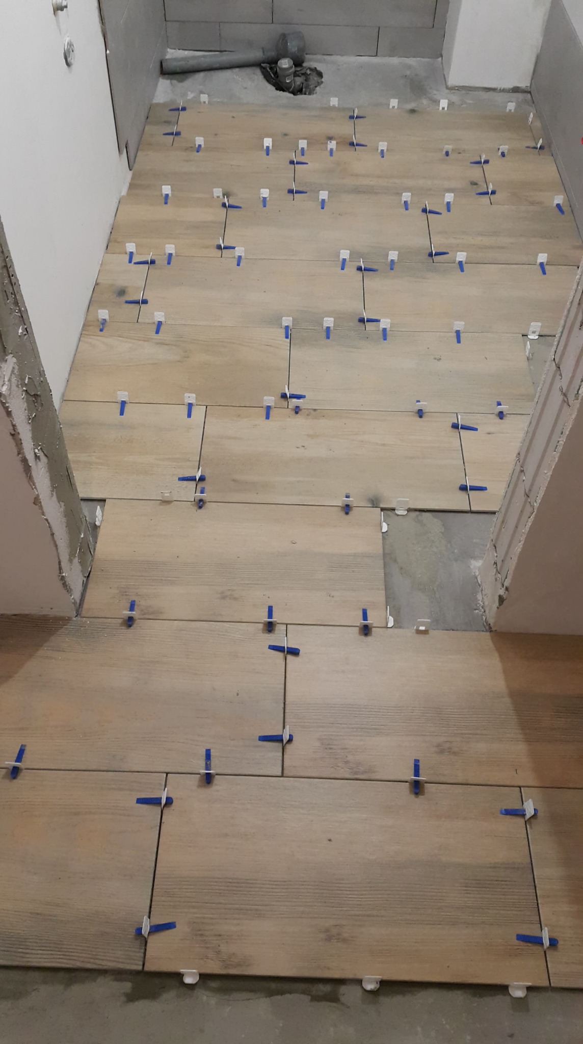 Układanie drewnopodobnych płytek podłogowych w łazience z użyciem niebieskich krzyżyków dystansowych, widoczne niedociągnięcia przy ścianie i brak fugowania.