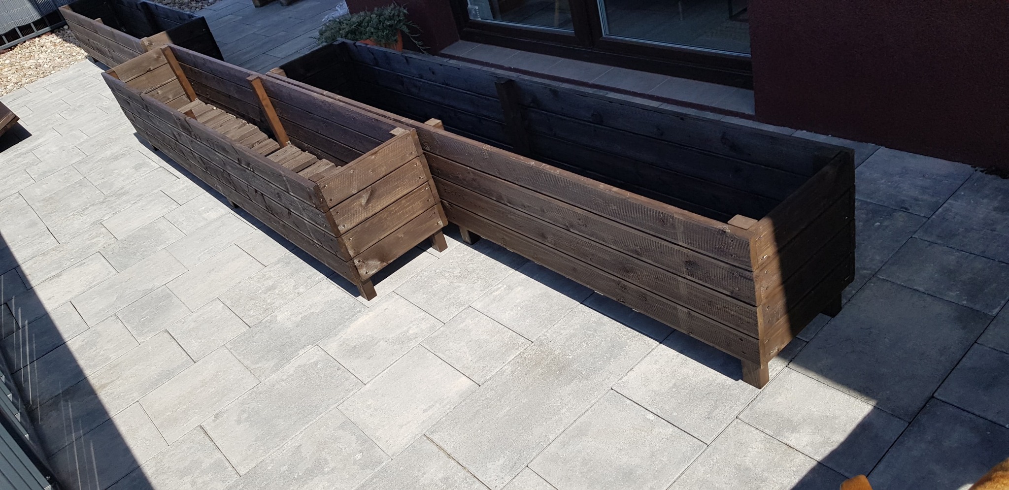 Dwa drewniane kwietniki ogrodowe w odcieniach brązu, ustawione na szarym bruku, z rośliną w doniczce w tle.