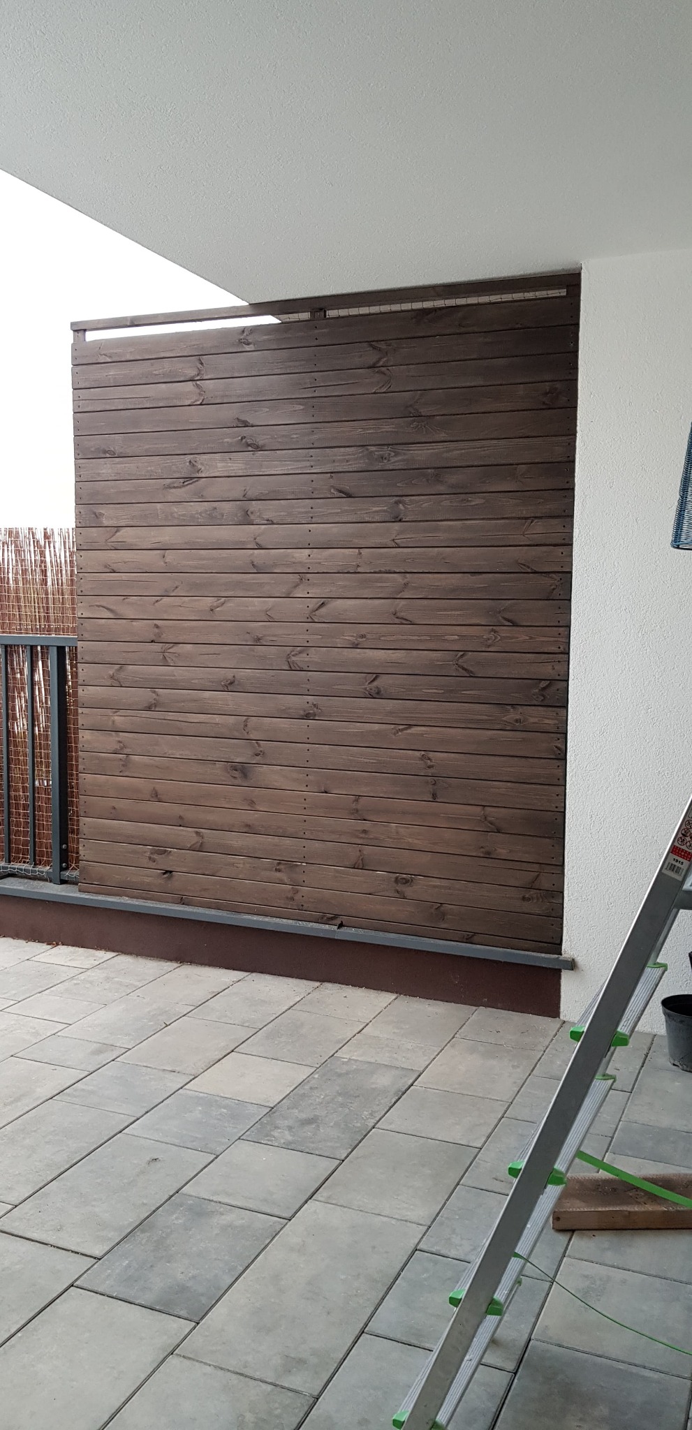 Drewniana, pozioma przesłona balkonowa w kolorze ciemnego brązu, zamontowana na szarym balkonie wyłożonym płytkami, widoczna drabina aluminiowa oparta o ścianę.