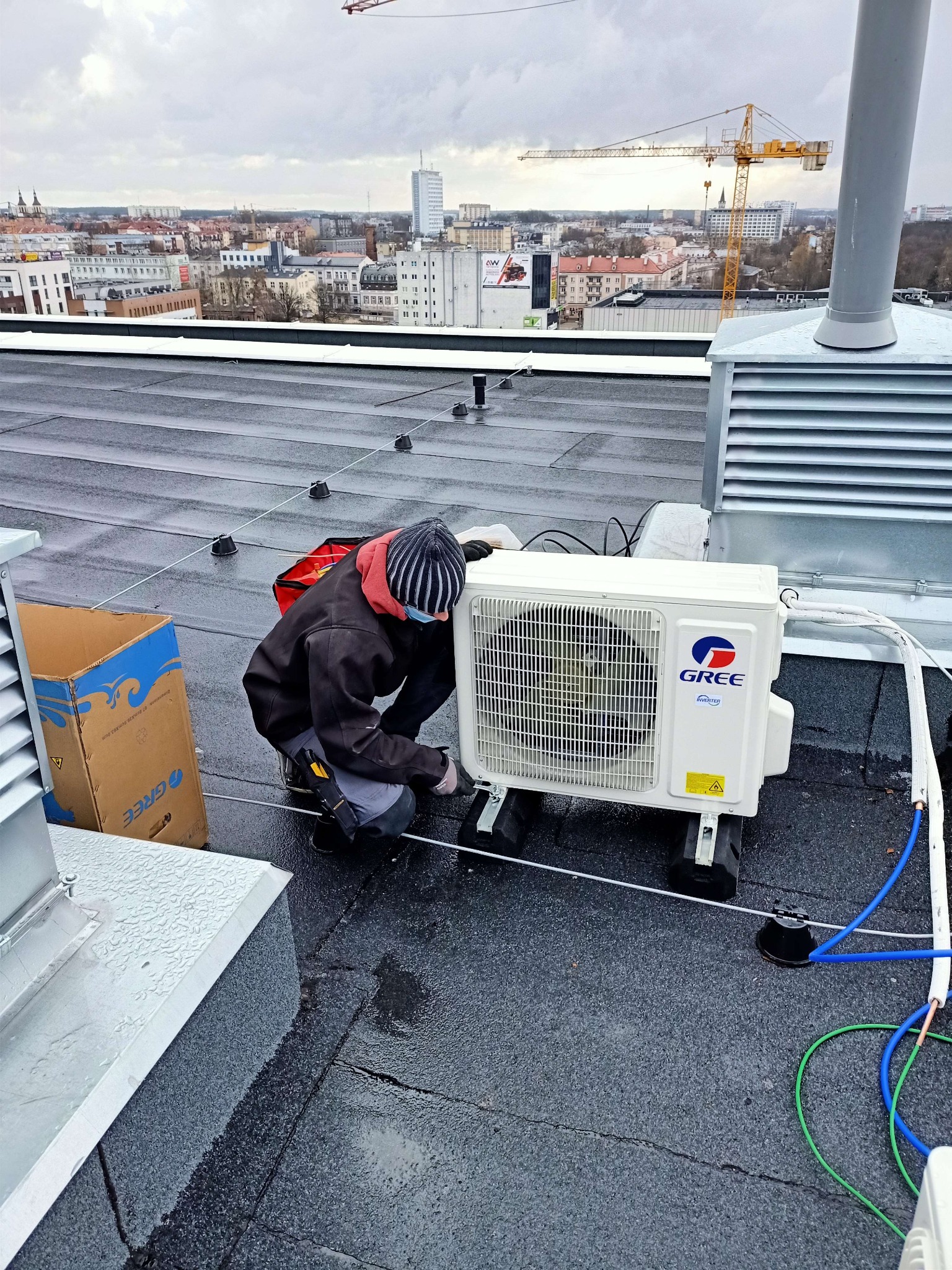 Montaż klimatyzatora marki Gree na dachu budynku w Białymstoku, widoczna panorama miasta i dźwig budowlany w tle.