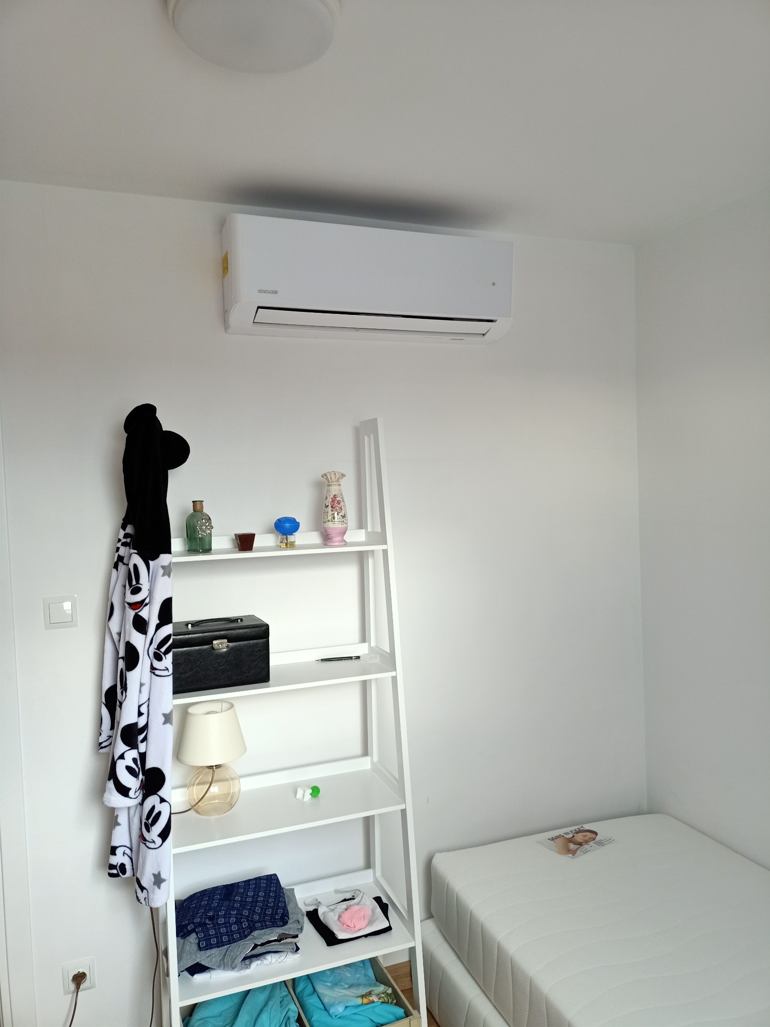 Biały klimatyzator Sinclair zamontowany na ścianie nad białym regałem drabinkowym w jasnym pokoju z łóżkiem w rogu.