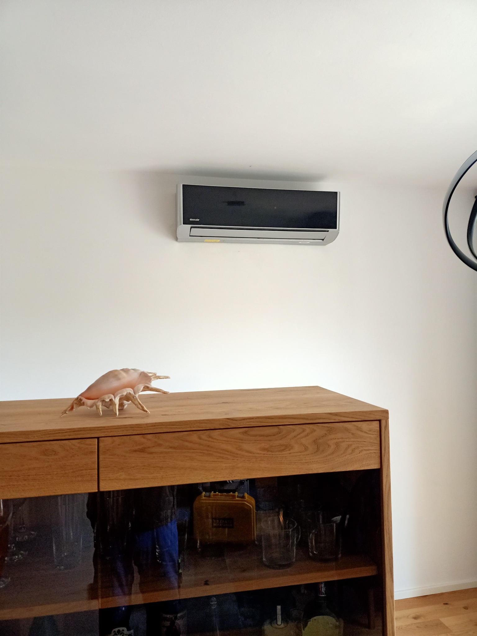 Zamontowany klimatyzator Sinclair na białej ścianie nad drewnianą komodą z dekoracyjną muszlą
