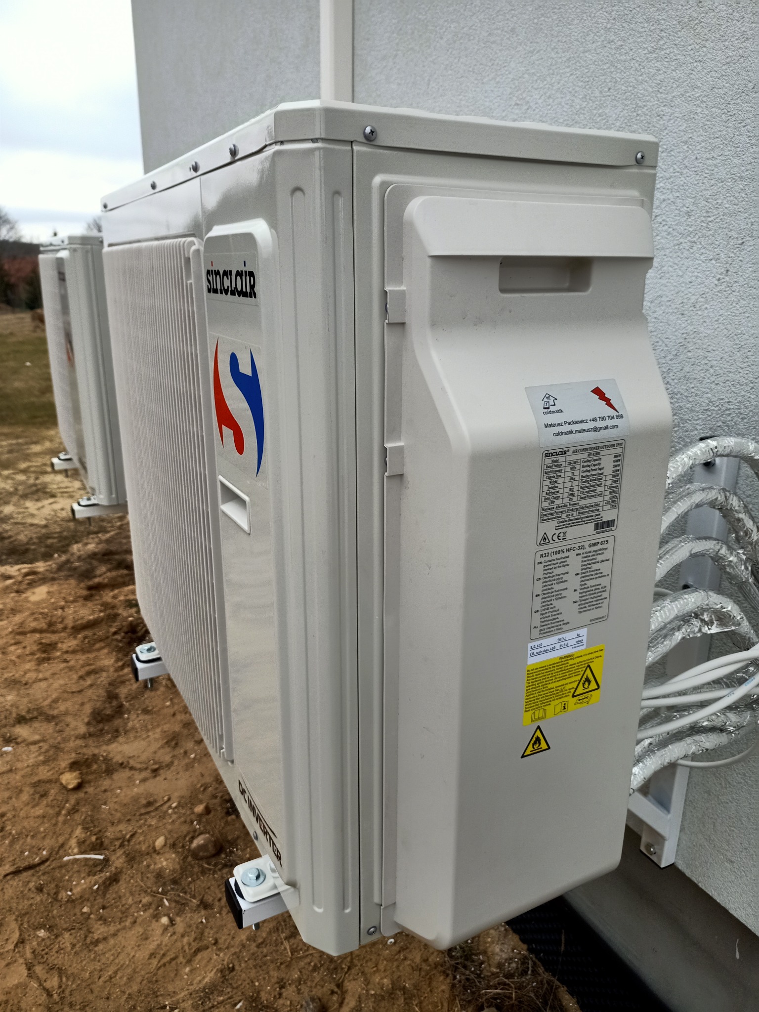 Zewnętrzne jednostki klimatyzacji Sinclair zamontowane na elewacji budynku, widoczne przewody w srebrnej izolacji oraz tabliczka znamionowa z danymi technicznymi.