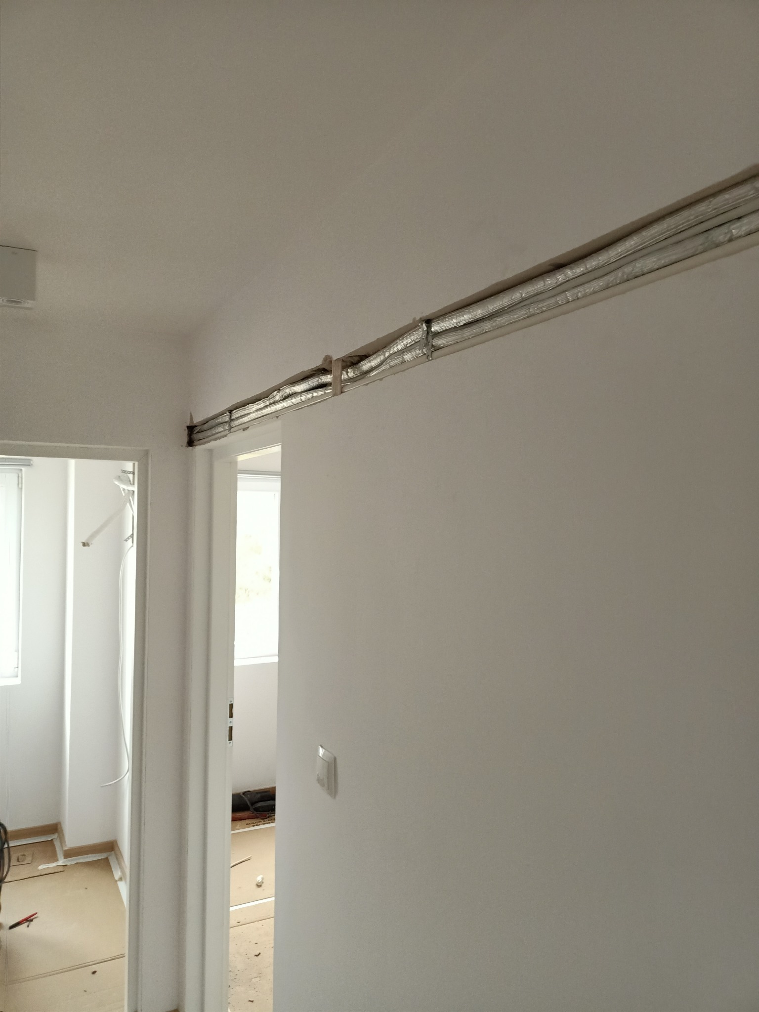 Montaż przewodów wentylacyjnych w nowym budynku, widoczne izolowane rury pod sufitem i fragment instalacji w przejściu między pomieszczeniami.