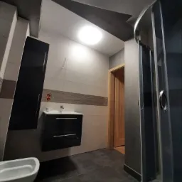 Nowoczesna łazienka z szarymi płytkami podłogowymi, beżowymi płytkami ściennymi z dekoracyjnym pasem, czarnymi szafkami wiszącymi, białą umywalką i toaletą oraz kabiną prysznicową z grafitowymi...