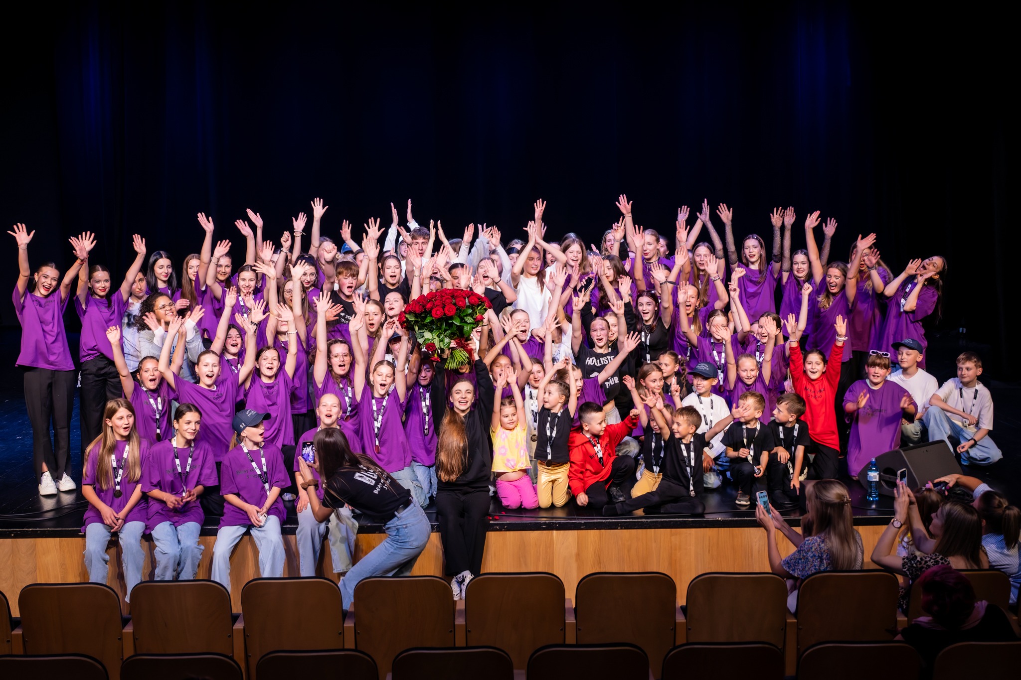 Duża grupa dzieci i młodzieży w fioletowych koszulkach z podniesionymi rękami na scenie, w centrum instruktorka z bukietem czerwonych róż, widownia z przodu.