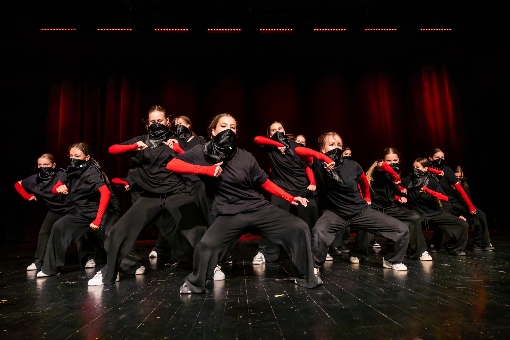 Grupa tancerzy w czarnych strojach i czerwonych rękawach wykonuje synchroniczny układ na scenie z drewnianą podłogą, podświetlona czerwonymi światłami.