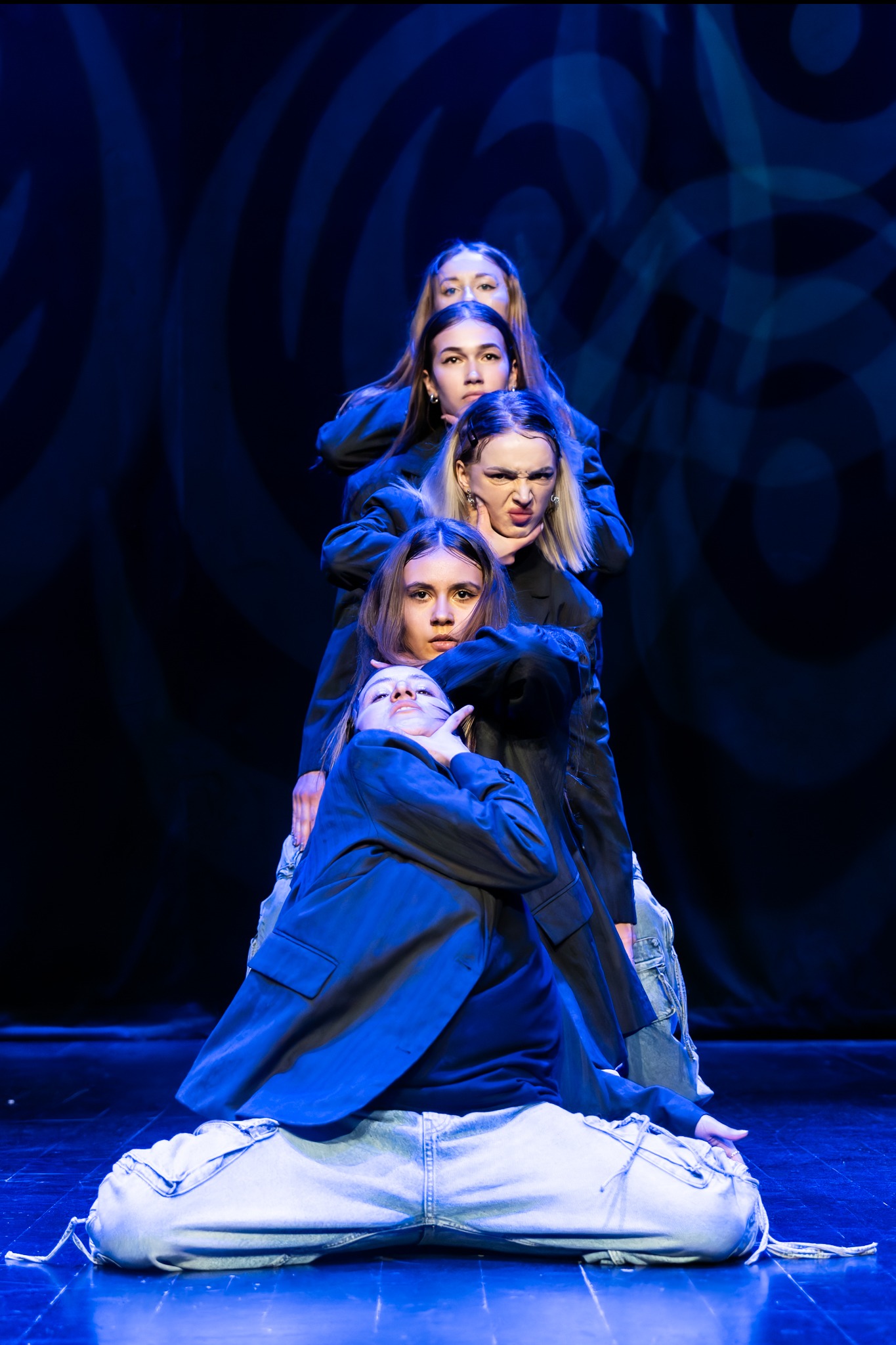 Cztery tancerki w dynamicznej pozie na scenie w niebieskim oświetleniu, ubrane w jeansy i marynarki, tworzące piramidę ciał.