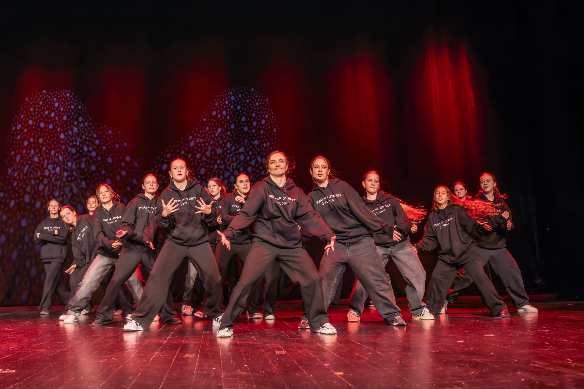 Zespół nastolatek w czarnych bluzach z kapturem, z logo 'Beat of Street Studio' na scenie z czerwonymi światłami i wzorzystym tłem, wykonujących zsynchronizowany układ taneczny.