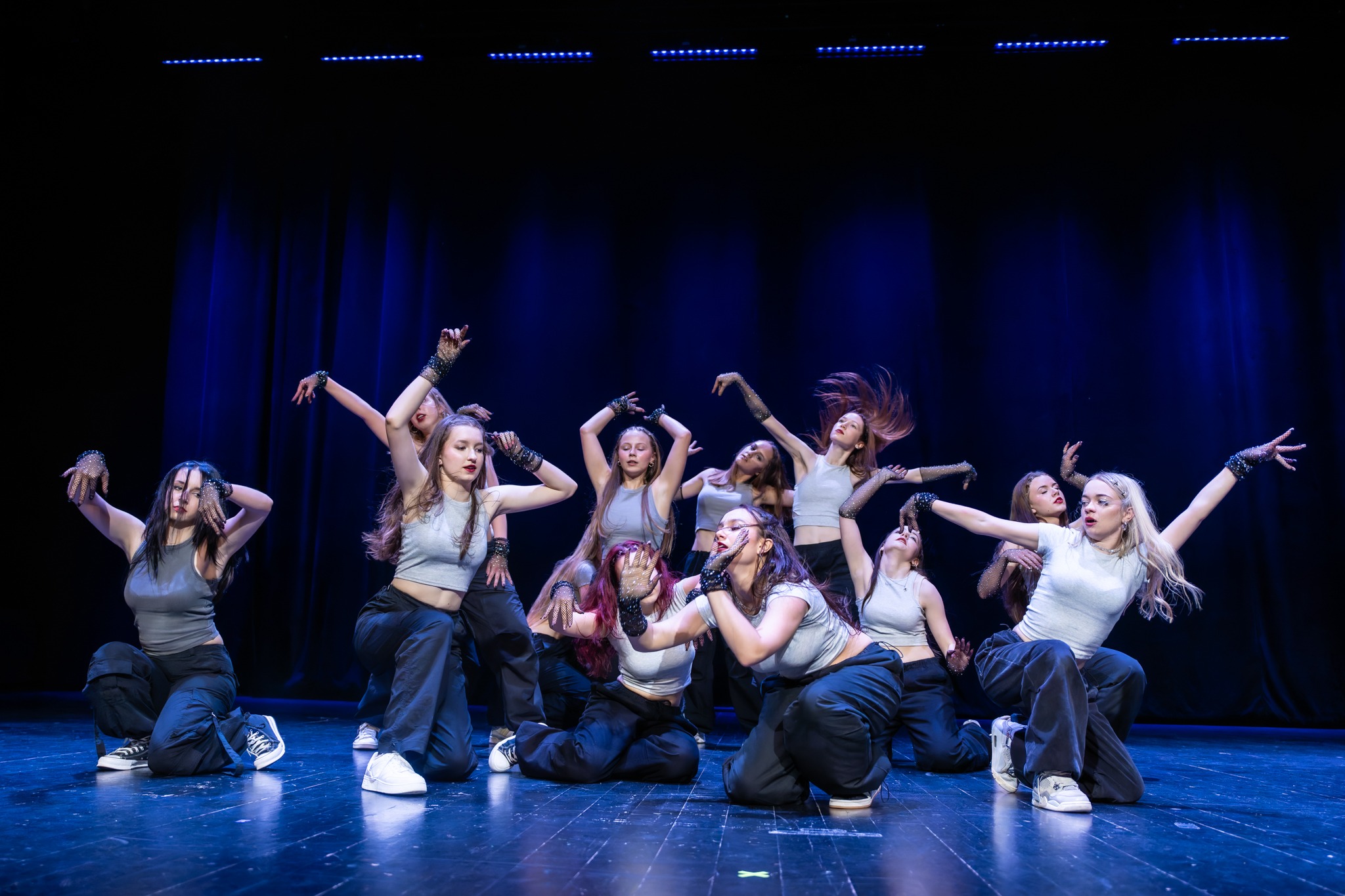 Grupa tancerek w szarych topach i czarnych spodniach cargo prezentuje choreografię na scenie z niebieskim oświetleniem i czarną kurtyną w tle, widać błyszczące rękawiczki.