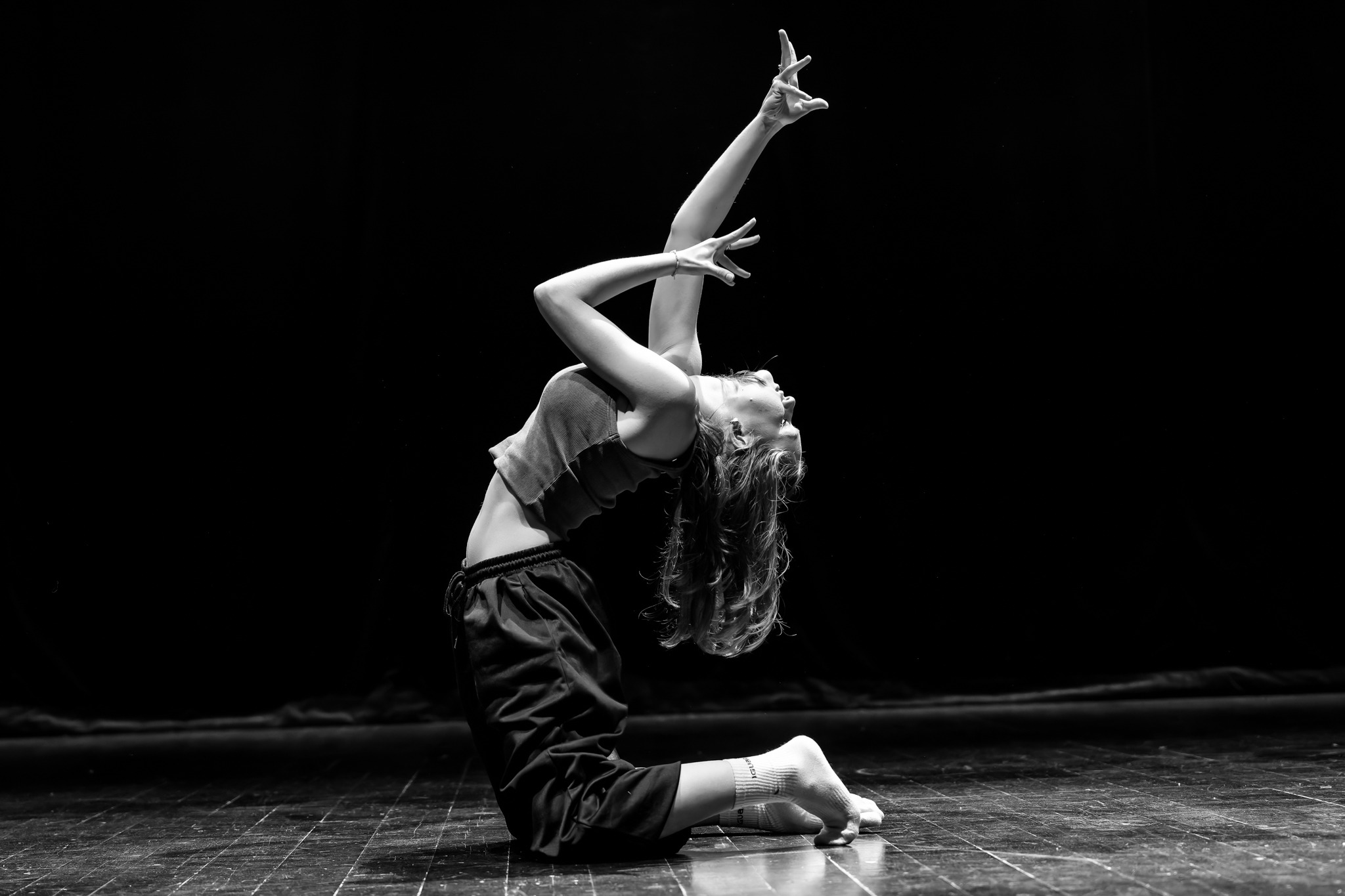 Czarno-białe zdjęcie kobiety w sportowym stroju, wykonującej wygięcie do tyłu na parkiecie z podniesionymi rękoma, na tle czarnego tła.