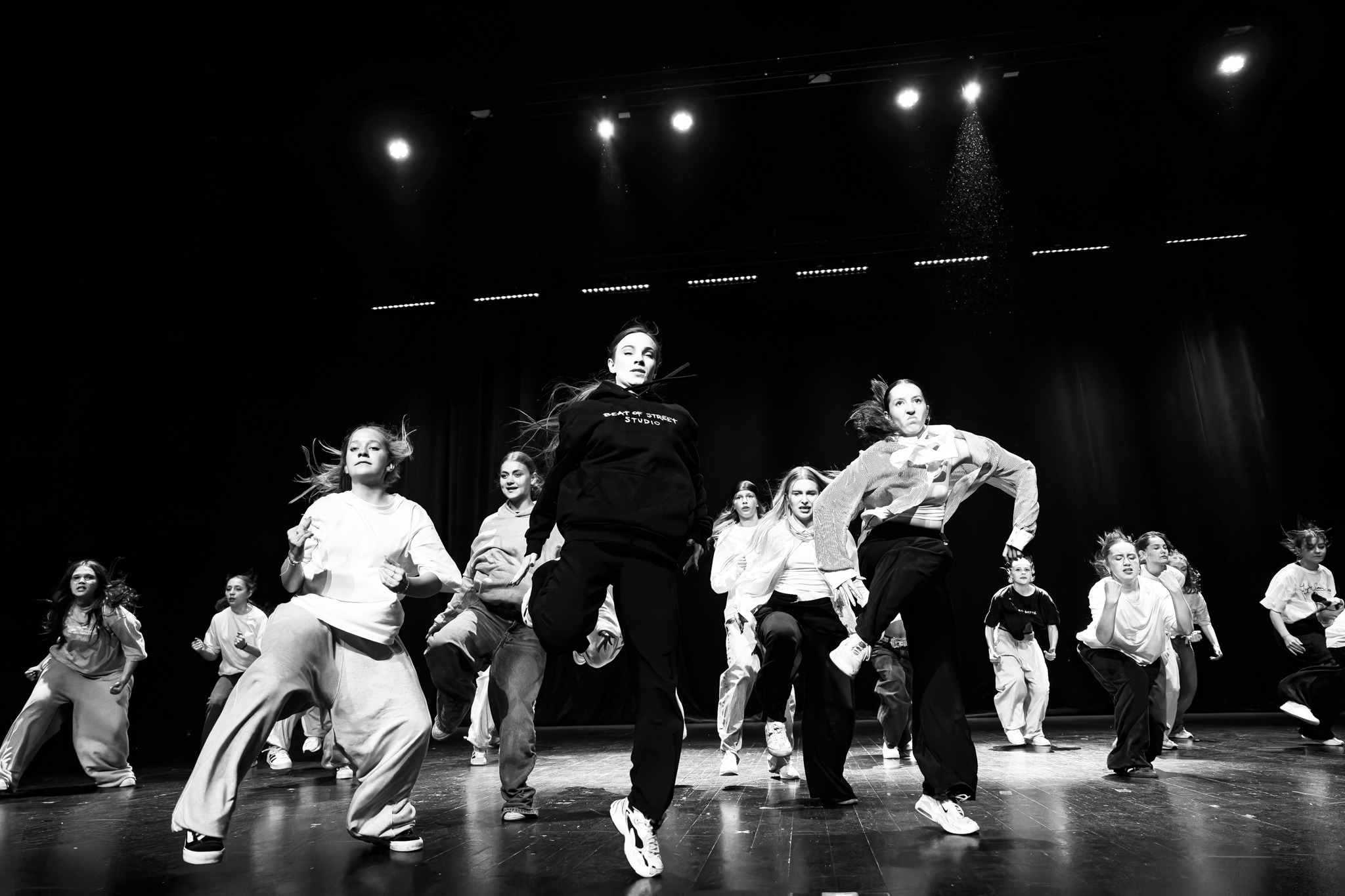 Czarno-białe zdjęcie grupy nastolatków wykonujących dynamiczny układ taneczny na scenie z ciemnym tłem i padającym światłem punktowym, jedna z tancerek ma na sobie bluzę z napisem 'BEAT OF STREET...