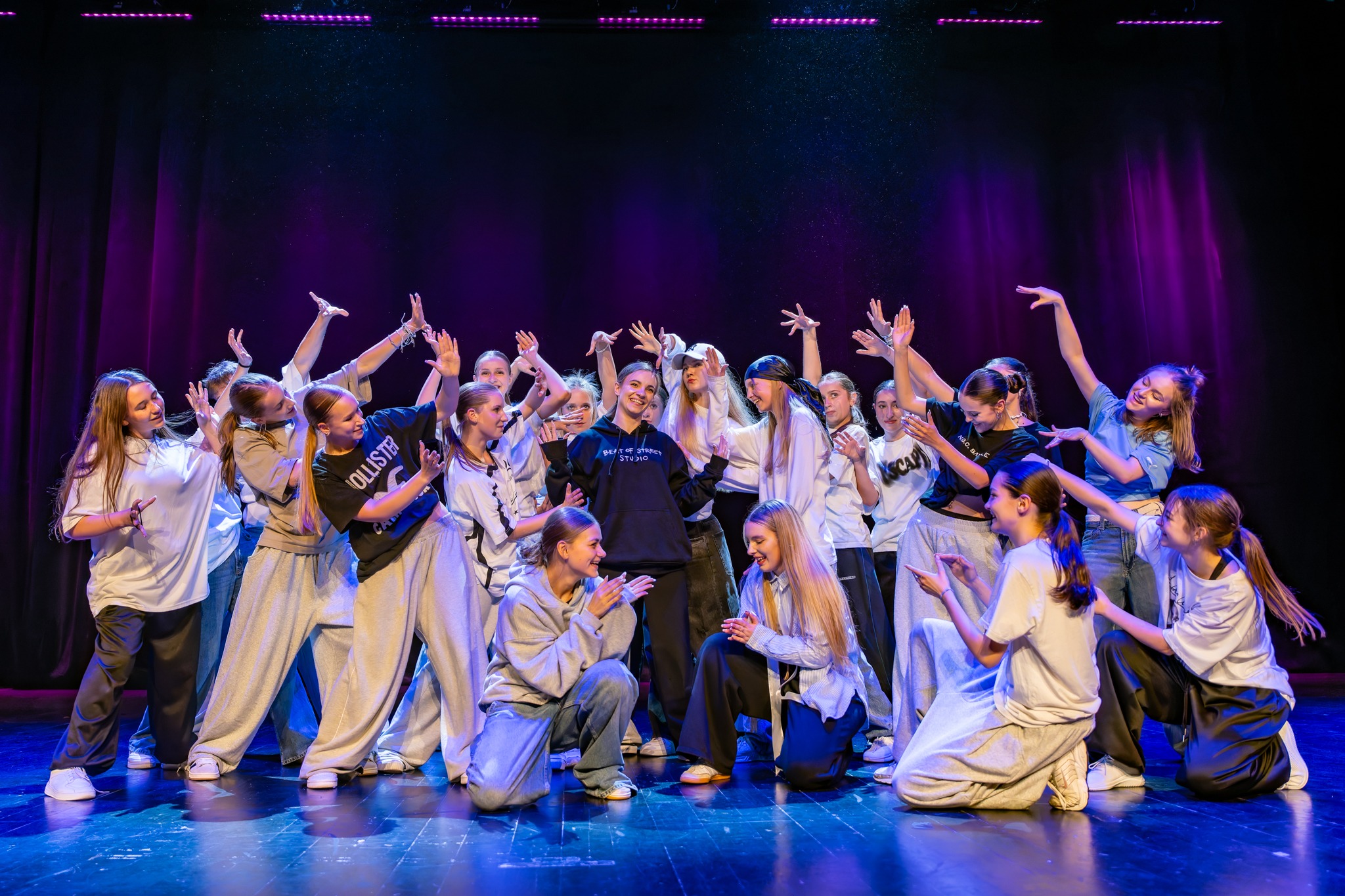 Grupa młodych tancerzy w luźnych strojach pozuje na scenie z fioletowym oświetleniem, liderka w czarnej bluzie z napisem 'Beat of Street Studio' w centrum.