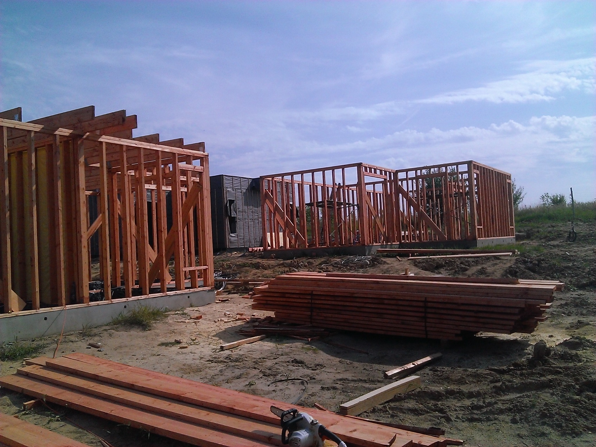 Konstrukcja szkieletowa drewnianych domów w trakcie budowy, widoczne fundamenty, stos drewna i piła łańcuchowa na placu budowy w słoneczny dzień.