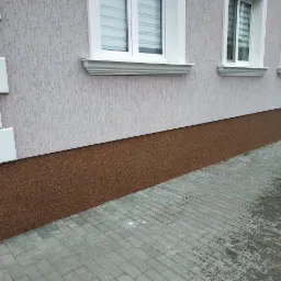 Wykończona elewacja domu jednorodzinnego z jasnoszarym tynkiem strukturalnym powyżej i ciemnobrązowym cokołem z żywicy poniżej, widoczne białe obramowania okien i chodnik z szarej kostki brukowej.