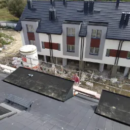 Montaż ocieplenia elewacji budynku wielorodzinnego w Krakowie, widok z góry na rusztowanie, na którym pracują dwie osoby, w tle budynek w trakcie prac wykończeniowych, na dachu widoczny fragment...