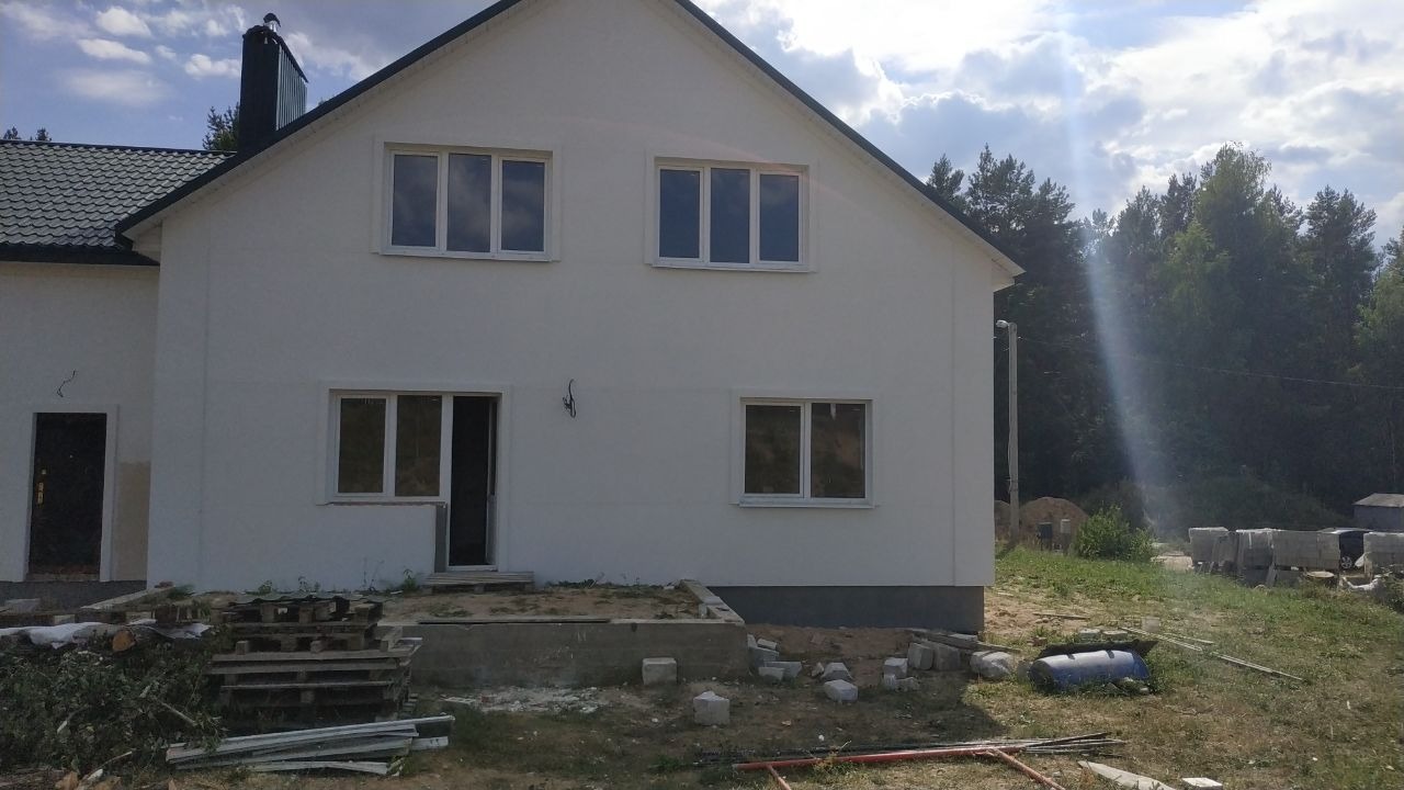 Biały dom jednorodzinny z ciemną dachówką, widoczne okna, drzwi wejściowe, fragment elewacji i teren wokół domu w trakcie prac wykończeniowych.