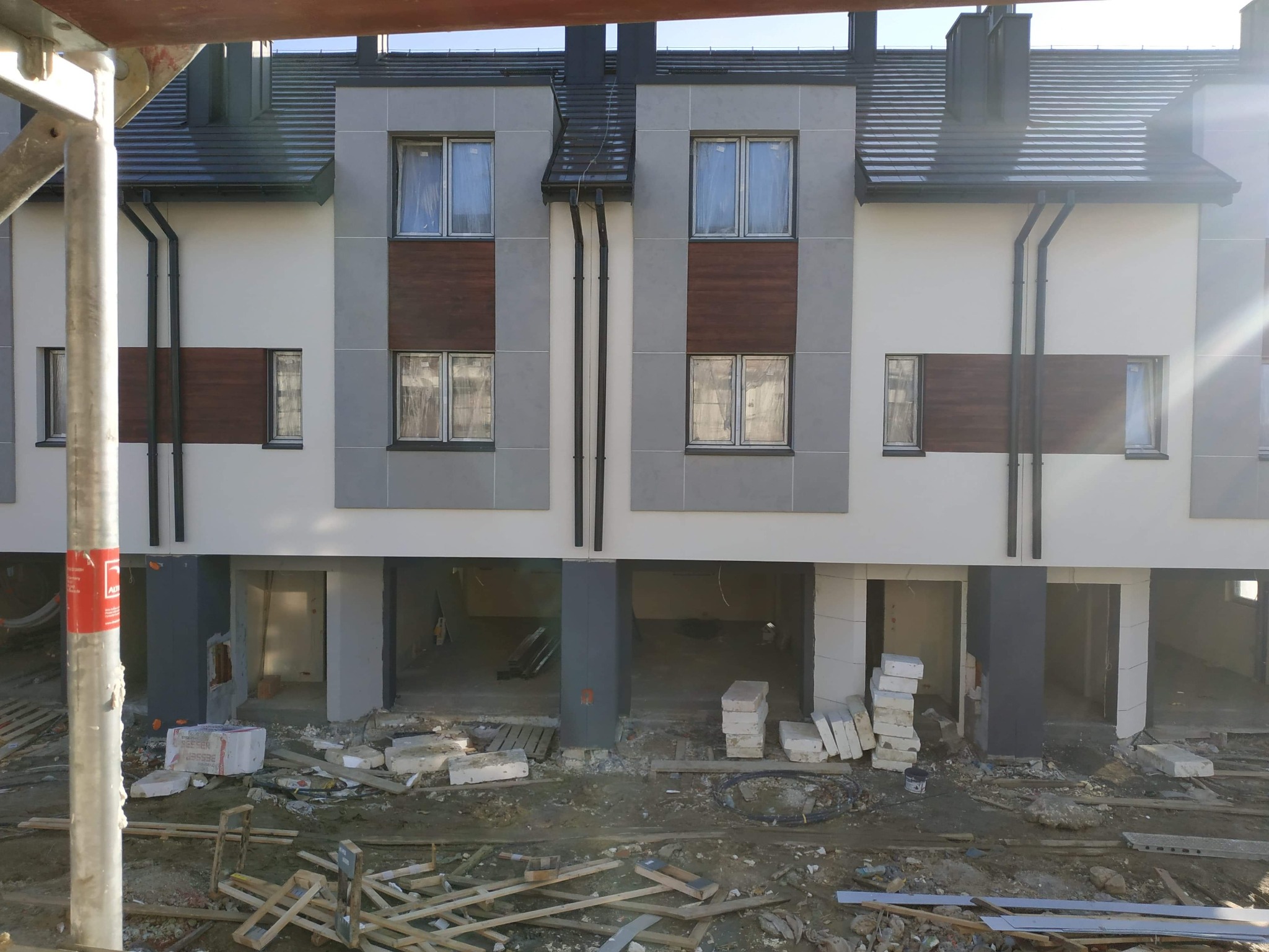 Surowy stan elewacji szeregowca z widocznymi elementami konstrukcyjnymi i stertami materiałów budowlanych na placu budowy.