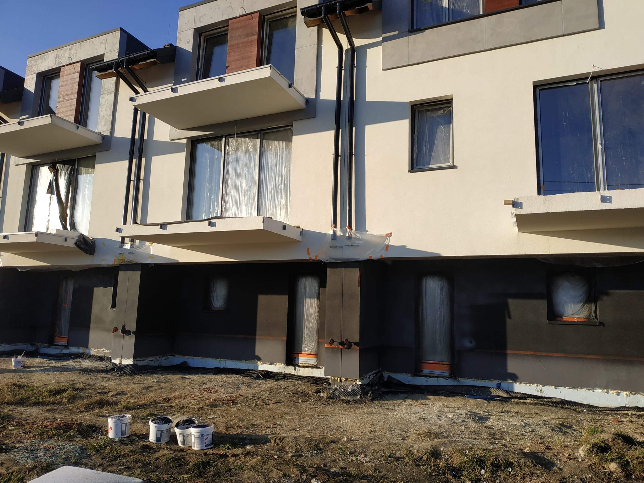 Fragment elewacji budynku wielorodzinnego w trakcie prac ociepleniowych; widoczne zabezpieczone okna folią, częściowo ocieplona dolna partia ściany i puste wiadra po farbie na ziemi.