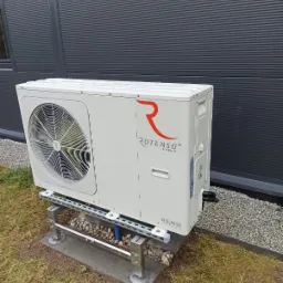 Biała pompa ciepła Rotenso Aquami Series zamontowana na metalowej konstrukcji na betonowej podstawie, z widoczną szarą elewacją budynku w tle.