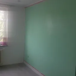 Pokój po częściowym pomalowaniu na zielono, z taśmą malarską zabezpieczającą krawędzie, grzejnik pod oknem.