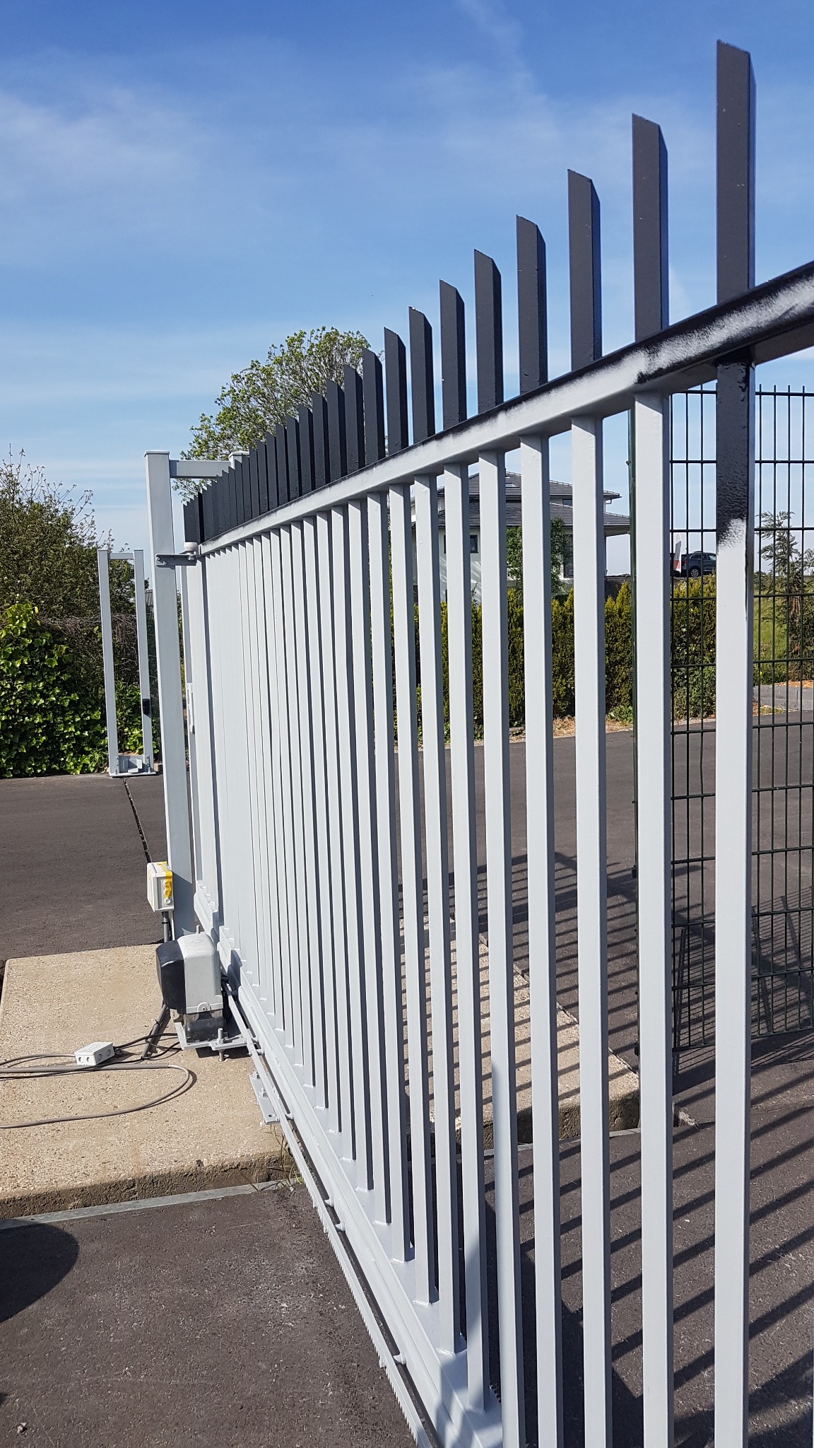 Automatyczna brama przesuwna z szarymi pionowymi prętami i czarnymi, ukośnie ściętymi elementami na górze, widoczny mechanizm napędowy i betonowa podstawa.