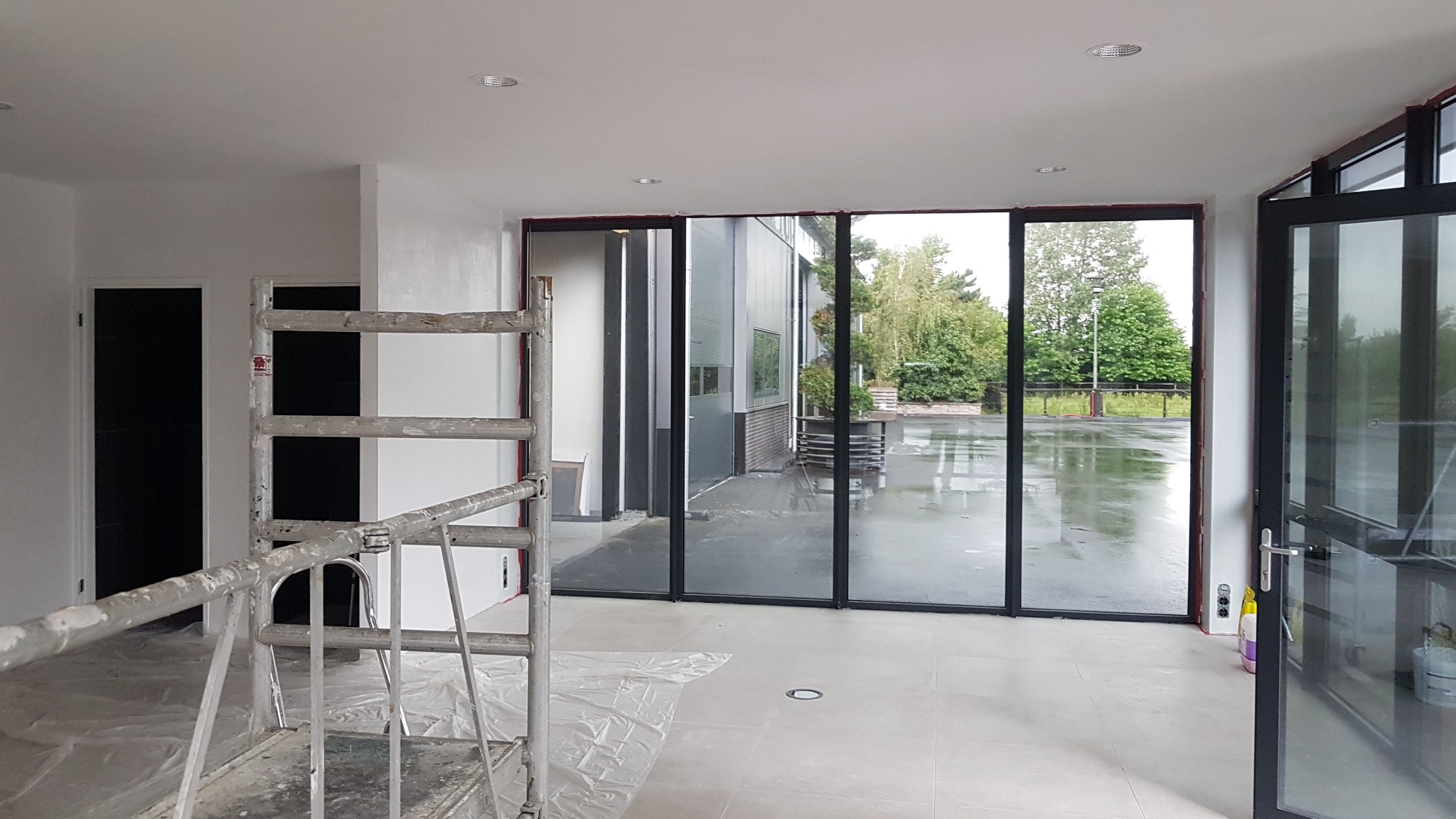 Wnętrze w trakcie malowania: białe ściany, drabina malarska z widocznymi śladami farby, podłoga zabezpieczona folią malarską, duże okna z widokiem na zewnątrz w pochmurny dzień.