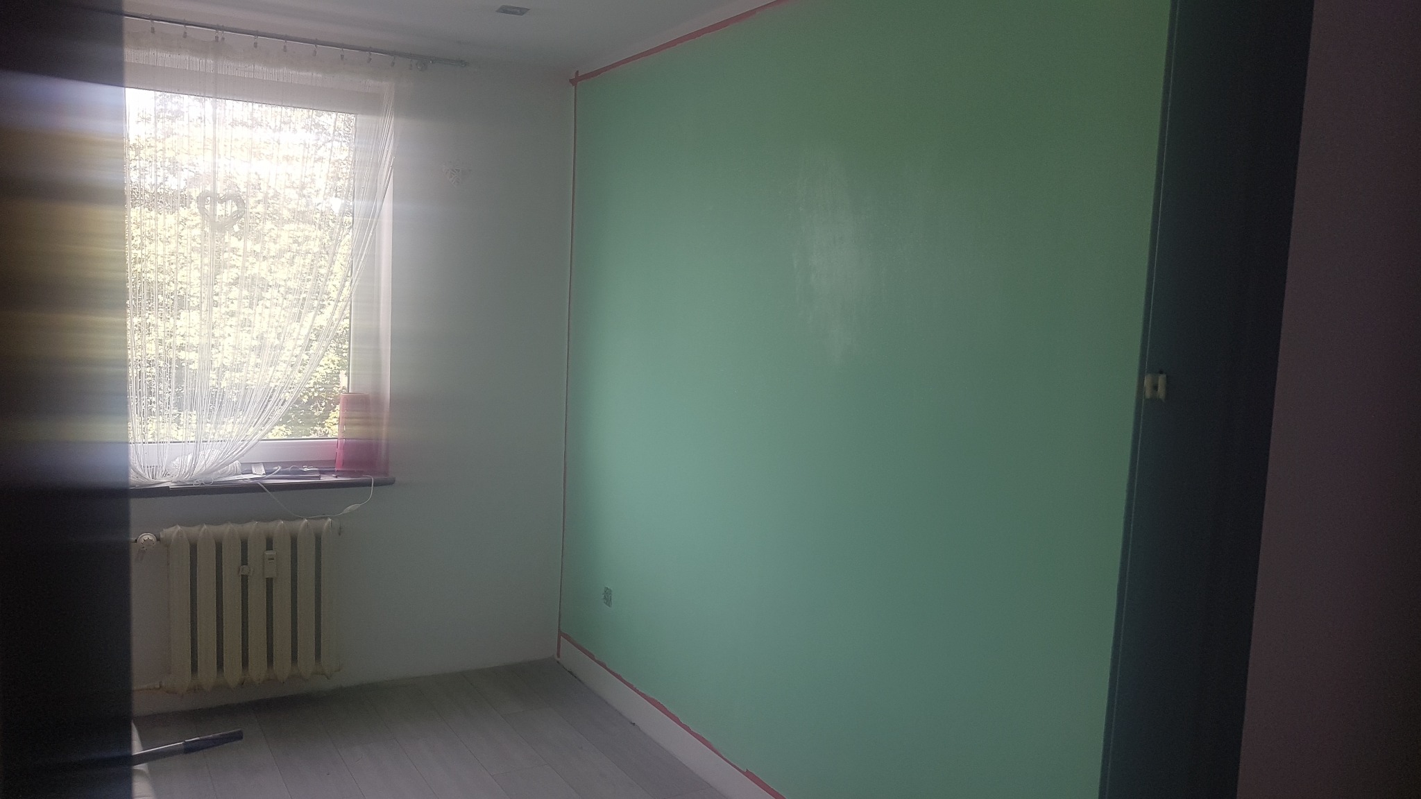 Pokój po częściowym pomalowaniu na zielono, z taśmą malarską zabezpieczającą krawędzie, grzejnik pod oknem.