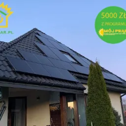 Dom jednorodzinny z czarnym dachem pokrytym panelami słonecznymi; widoczny baner informujący o dotacji 5000 zł z programu Mój Prąd.