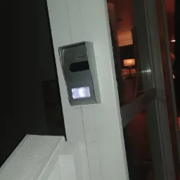 Zewnętrzny panel wideodomofonu z podświetleniem LED zamontowany na białej futrynie drzwi wejściowych, widok na wnętrze domu z oświetloną lampą podłogową w tle.