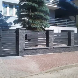 Nowoczesne ogrodzenie z poziomych, szarych paneli i słupków z szarego kamienia łupanego, brama wjazdowa z tego samego materiału, na tle domu z szarą elewacją.