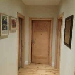 Korytarz po remoncie: Świeżo pomalowane ściany w odcieniach beżu, drewniana podłoga i drzwi z surowego drewna, dekoracyjne obrazy i lustro.