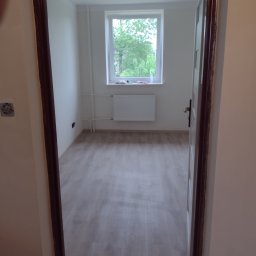 Prusik - Wykończone wnętrze pokoju z panelami podłogowymi w odcieniach szarości, widok przez otwarte drzwi z brązową ościeżnicą, okno z widokiem na zieleń na wprost, grzejnik pod oknem.