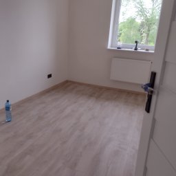 Prusik - Pomalowane na biało wnętrze pokoju z panelami podłogowymi, widoczna butelka wody na podłodze, okno z widokiem na zieleń i narzędzia na parapecie, oraz biały grzejnik.