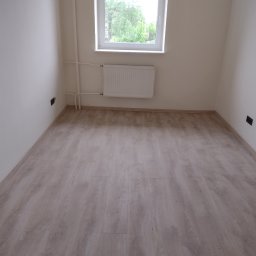 Prusik - Jasny pokój z nową podłogą z paneli imitujących drewno, biały grzejnik pod oknem, widoczne gniazdka elektryczne.