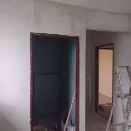 Wnętrze w trakcie remontu: ściany zagruntowane, widoczne ślady po gładzi, zamontowana płyta karton-gips w jednym z pomieszczeń, drabina malarska oparta o ścianę, widoczne narzędzia malarskie.