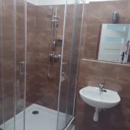 Błyszcząca kabina prysznicowa z brodzikiem, umywalka z baterią i lustro w brązowej łazience wyłożonej płytkami.