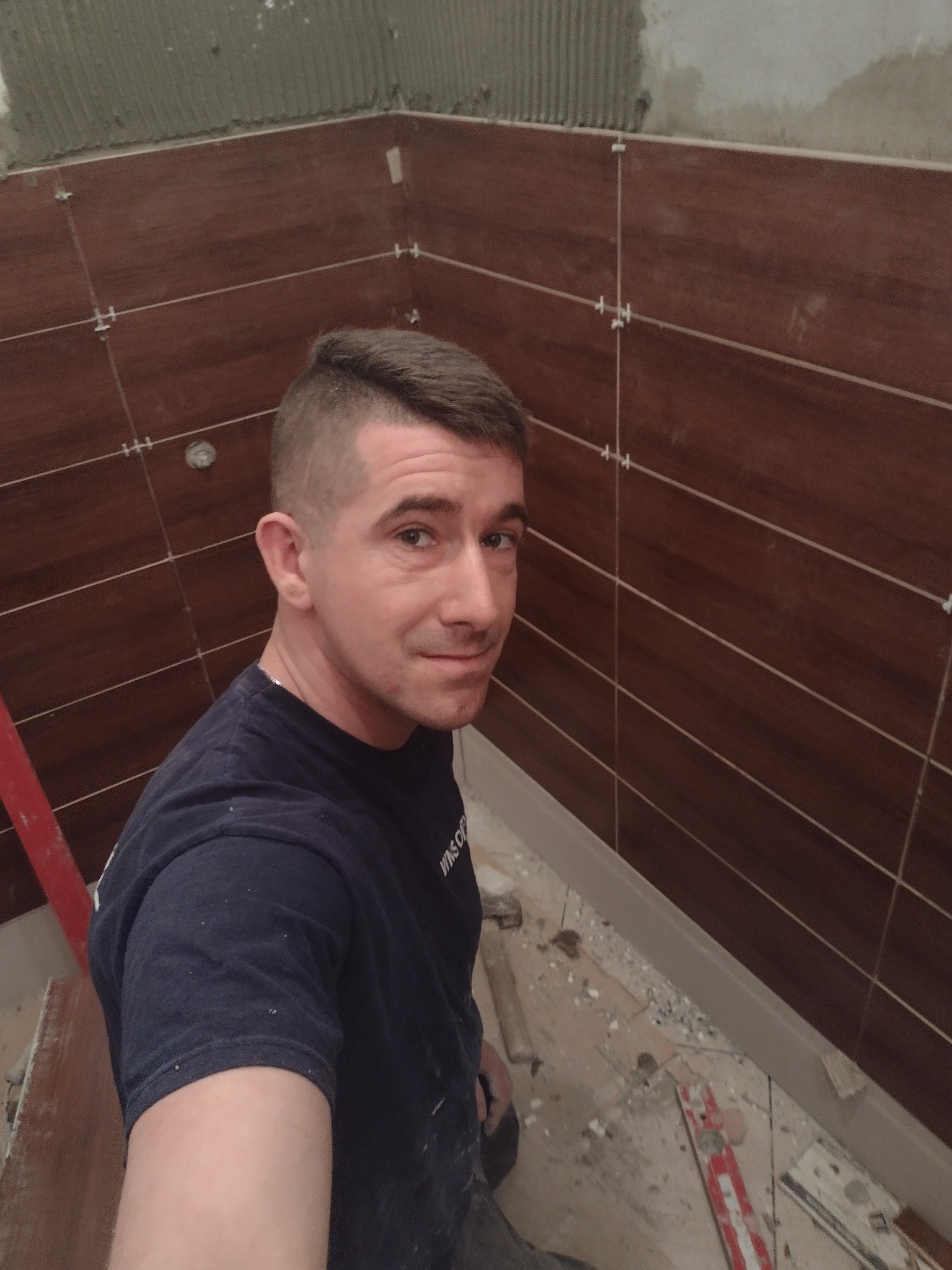 Mężczyzna uśmiecha się na tle brązowych płytek z białymi fugami w trakcie montażu. Widoczne krzyżyki dystansowe i fragment ściany z resztkami kleju. Na podłodze resztki materiałów budowlanych.